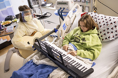 A healthcare worker playing a guitar and a child patient in a bed playing a keyboard.