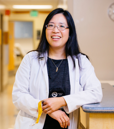 Elisa Tong smiles in a hallway while wearing a white medical coat over a black shirt.