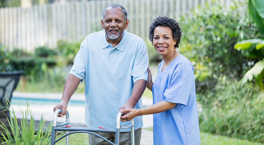 An older man using a mobility walker outside is assisted by a nurse. 