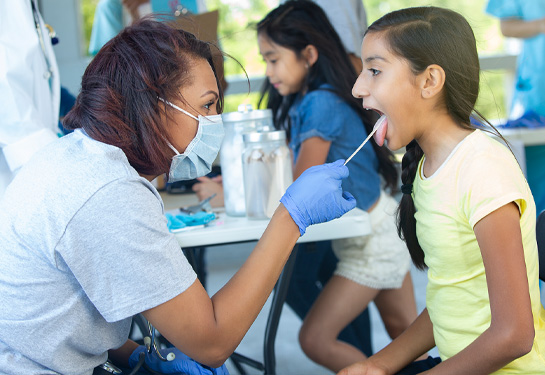 During a free clinic at the local park, a female clinician uses a tongue depressor to check preteen girl's throat. The clinician wears protective mask