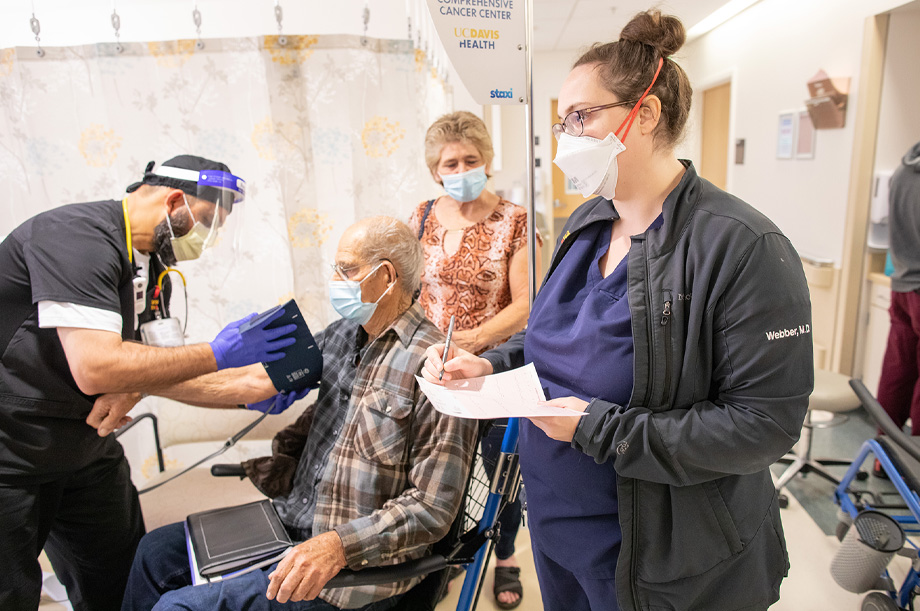 Nurse in ED places blood pressure cuff on geriatric patient who sits in a wheelchair while older woman holds chair handles and second nurse reviews paperwork; all wear masks