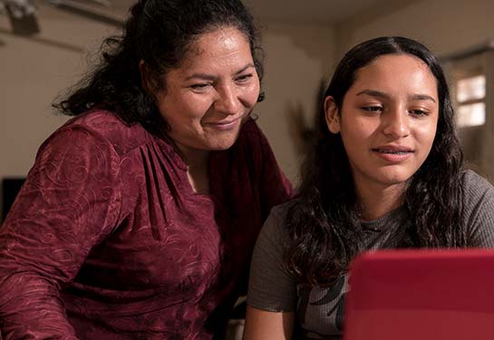 A woman and her teenage daughter stand before a computer screen – the woman smiles. 