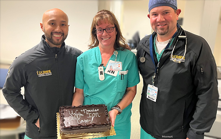Two men and a woman wearing scrubs hold a brown cake with white writing that states, 'UC Davis 100th Ion Case!'