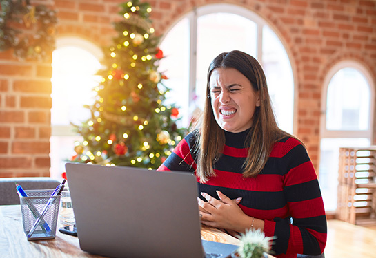 Woman sitting at the table working with laptop at home around Christmas tree with hand on stomach because indigestion.