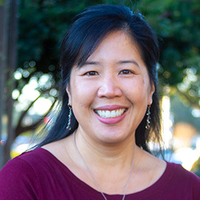 A headshot of a woman with black hair, wearing a burgundy shirt, smiling outside against a backdrop of trees. 