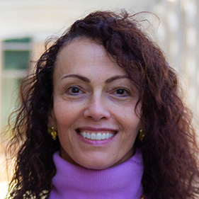 A headshot of a woman with red hair, wearing a light purple shirt, smiling outdoors. 