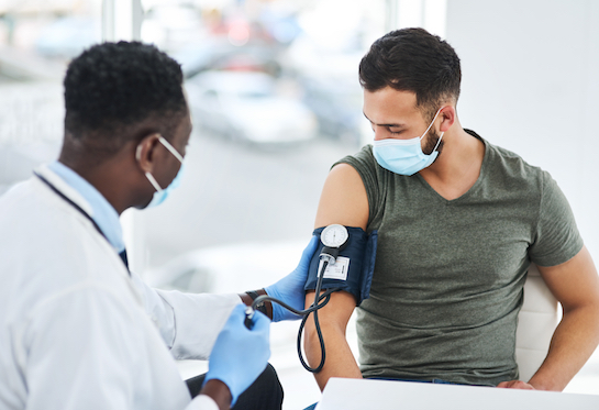 A doctor in a white coat and surgical mask holds a gloved hand on a blood pressure cuff that’s wrapped around the left arm of a young man