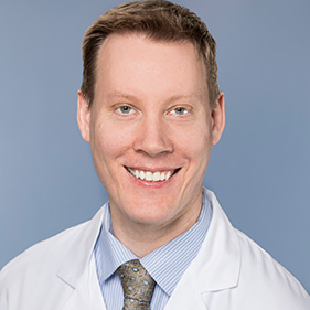 A man with short blonde hair wearing a white physician’s coat