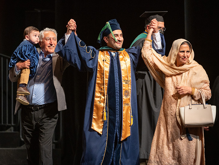 Graduating medical student in blue gown and yellow stole walks across stage to receive his diploma accompanied by family members 
