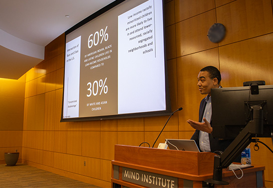 A man stands behind a wooden podium and in front of a wood-paneled wall. to his right is a large screen with a PowerPoint presentation projected