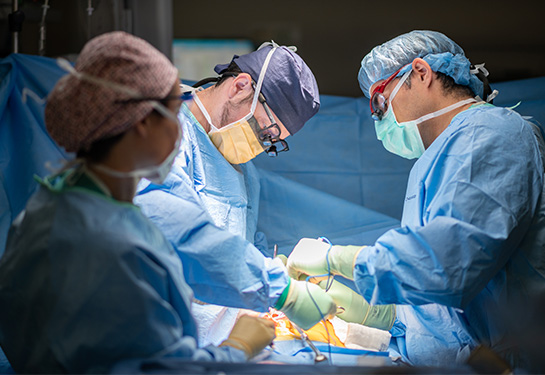 Three people dressed in blue scrubs standing over patient lying in bed.