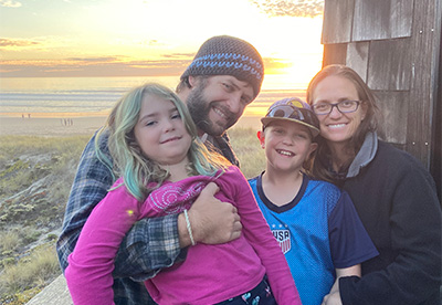 Family including adult woman and man posing with beach in background. Dad is holding small blonde girl smiling and her brother is smiling behind her mother.
