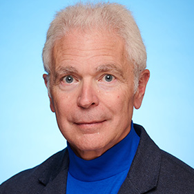 A man with white hair and wearing a dark sport coat against a light blue background.