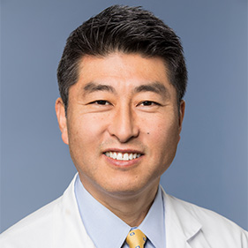 Surgeon Kee Kim, wearing a medical white coat, smiles inside. 