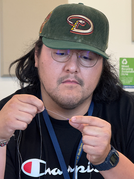 A young man in green baseball cap and black shirt stretches a thread with fingers from both hands to put beads through it
