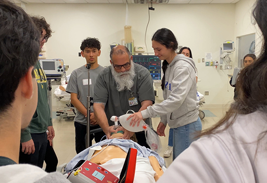 A man with a beard stands between high school students and teaches them to pump air into a training manikin&#x2019;s nose and mouth 