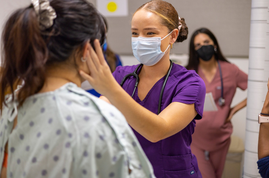A medical student in purple scrubs and surgical mask uses her left hand to examine a fellow student’s face in clinical practice