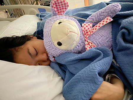 woman lying in hospital bed with blue blanket and stuffed animal. 