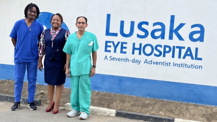 Two men and one woman stand in front of a white sign with blue lettering that says “Lusaka Eye Hospital.”