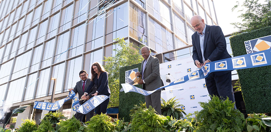 Four adults cut a ribbon on a stage in front of a large glass building. 