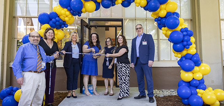 A group of people gather under a blue and gold balloon arch for a ribbon cutting