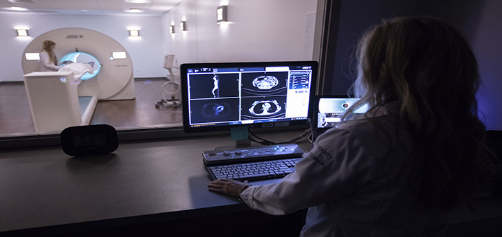 : Woman looking at monitor with images in front of room featuring large scanner in background.