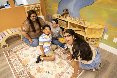 Woman playing with two boys and an older girl on a carpet surrounded by toys.