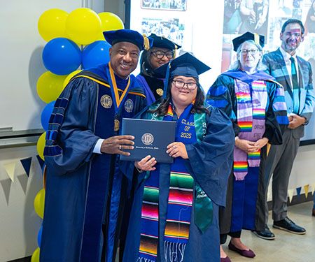 a scholar smiles with the chancellor and vice chancellor as she gets her diploma. 
