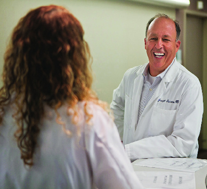 Man in white coat smiling at woman he is talking to across desk.
