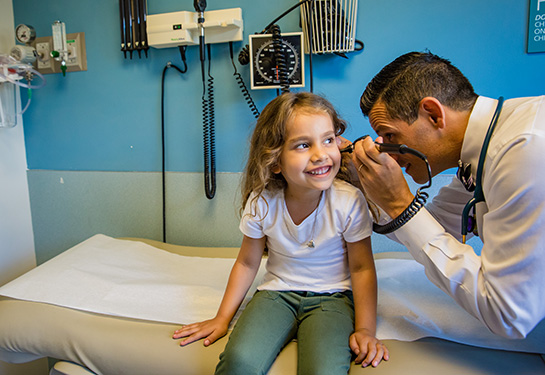 Doctor looks in girl&#x2019;s ear while she smiles on patient table.  