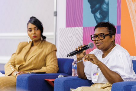 Woman holding mic sitting in blue chair while other woman looks on