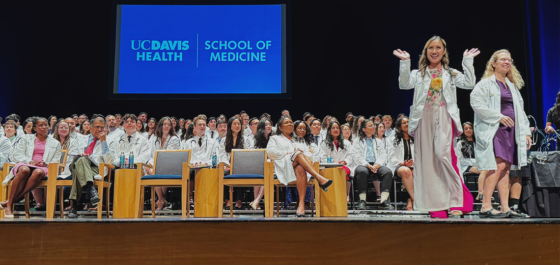 A medical student pumps her arms in celebration on stage at the white coat ceremony with fellow students seated in the background
