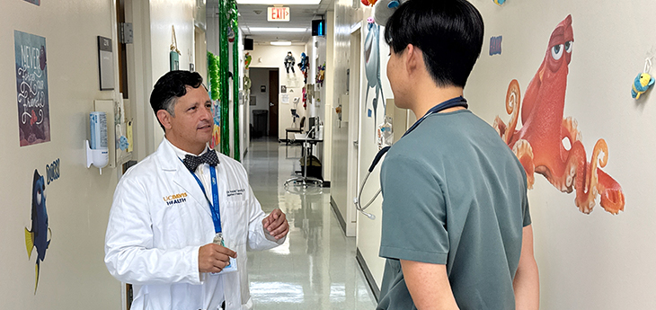 A doctor in a white coat uses hand gestures to instruct a medical student in scrubs in the hallway of a pediatric clinic.