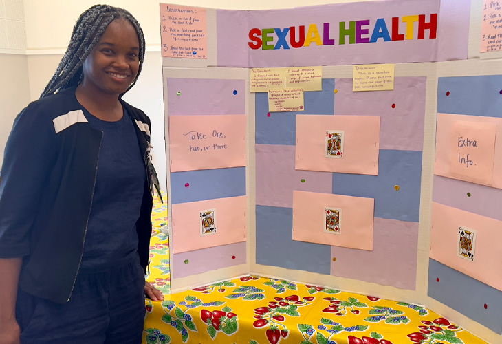 Nursing student in blue dress stands in front of colorful poster board presentation with title that reads “sexual health”