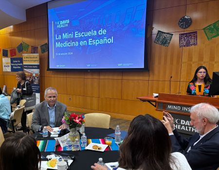 Participantes se sientan alrededor de distintas mesas en una sala donde una pantalla de proyección muestra una diapositiva que dice, “La Mini Escuela de Medicina en Español”