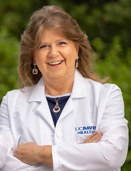 A woman wearing a white coat smiles with arms crossed, standing outdoors in front of greenery.