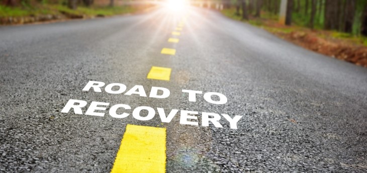 A paved road with yellow lines in a forest with the words ROAD TO RECOVERY.