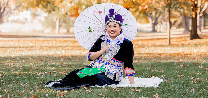 Smiling woman in traditional Hmong clothing sits on a blanket over grass. She is holding a parasol decorated with floral designs