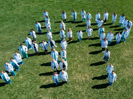 Health care professionals wearing white coats stand on a grassy field to spell out the letters A P P