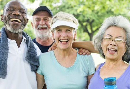 Four older adults stand outdoors smiling and laughing together after exercise, dressed in casual athletic clothing.