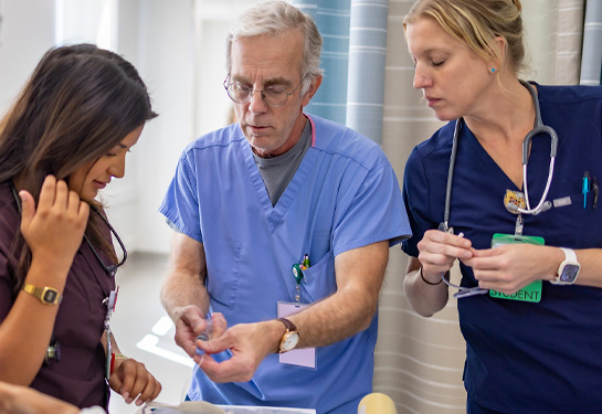 An instructor in blue scrubs demonstrates a procedure to two students who watch closely during a hands-on training session.