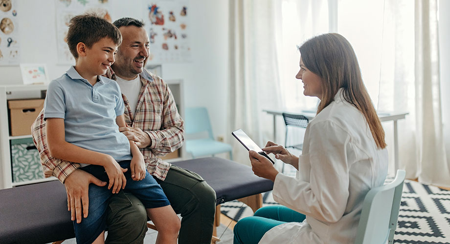 A boy sits on his father's lap in a medical office as a doctor sits across from them in a white coat.