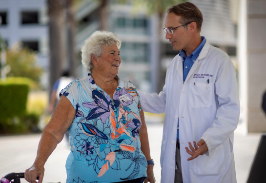 A person in a white coat speaking with someone in a floral shirt using a walker.