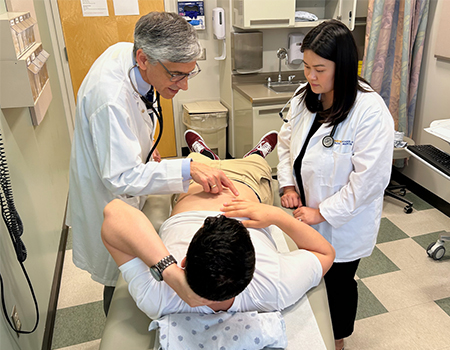 A doctor teaches a medical student how to examine a patient lying on an exam table in a clinic room during a training session