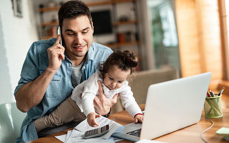 dad holding baby while on a phone call and trying to work on his laptop