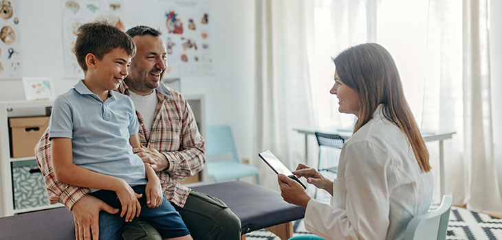 A man sits with his son on his lap while the two talk with a physician in a medical office.