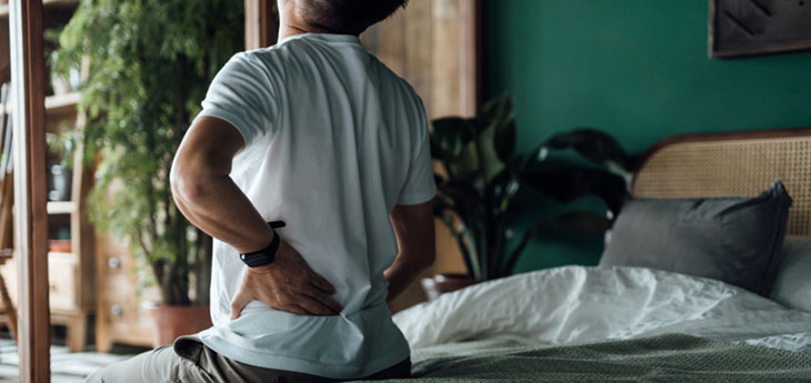 Rear view of senior Asian man suffering from backache, massaging aching muscles while sitting on bed.