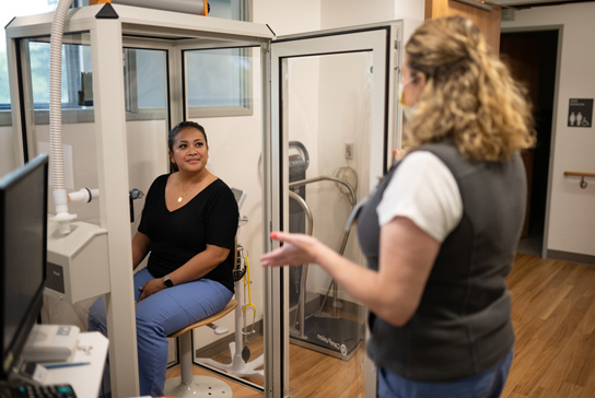 Woman in a lung capacity test chamber, speaking to a technician.